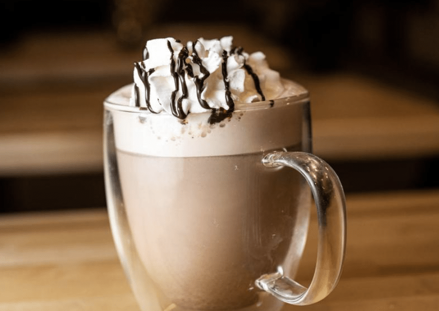 Glass of hot cocoa in a glass mug with whipped cream and a drizzle of chocolate syrup.